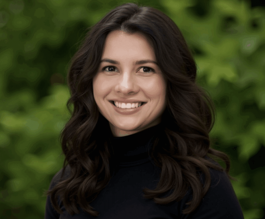 Smiling woman with long dark hair in black top, standing outdoors in front of green leafy background.